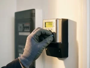 Gehandschoende elektricien die een slimme energiemeter instelt op witte muur, met consumentenunit op achtergrond.