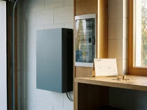 Thuisbatterij op witte garagemuur naast elektrisch paneel, met kalender en euromunten op plank die terugverdientijd symboliseren.